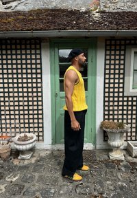 Suéter amarillo de punto con textura acanalada, pantalones negros de pierna ancha y zapatos amarillos con detalles verdes, apoyado contra una puerta verde.