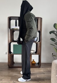 Personne en veste matelassée verte, pantalon noir, baskets blanches et écharpe noire couvrant la tête, debout de profil dans une pièce moderne avec une étagère en bois et une plante.