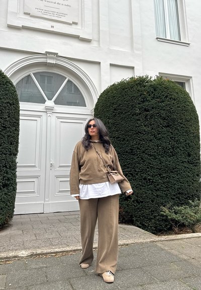 Mujer con pantalones holgados beige y sudadera sobre una camisa blanca, lleva gafas de sol y un bolso cruzado, de pie junto a grandes arbustos recortados.