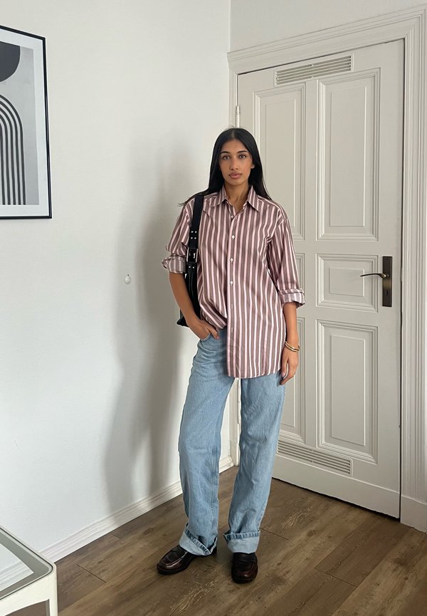 Randig skjorta i nyanser av rosa och vitt; tillsammans med ljusblå lösa jeans och mörkbruna skor. Svarta handväskedetaljer synliga.
