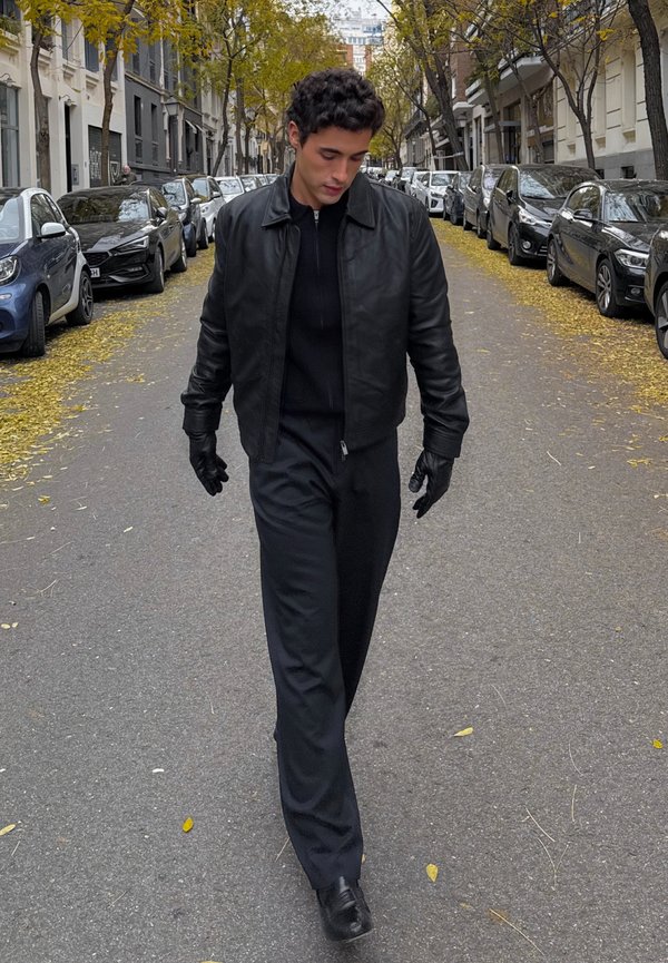 Man in black leather jacket, gloves, and pants walking down a tree-lined city street with parked cars and fallen yellow leaves.