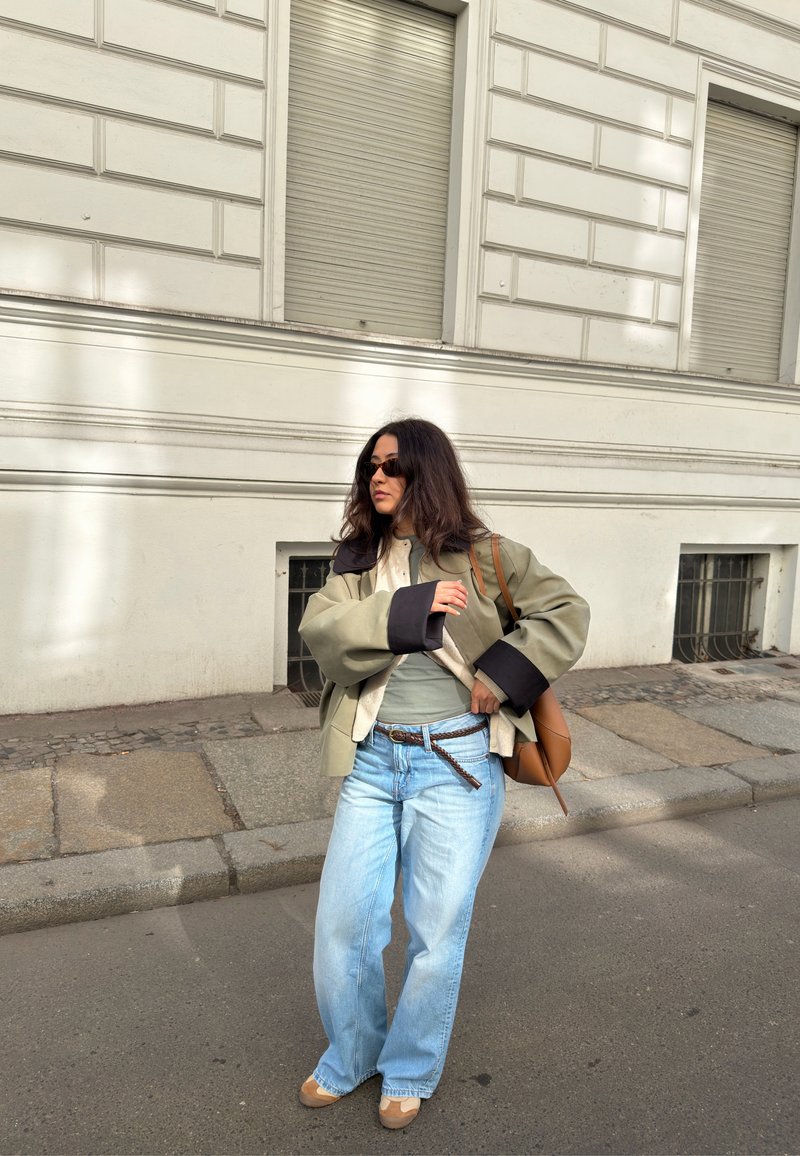 Femme aux longs cheveux foncés portant une veste oversize, un jean bleu clair, des lunettes de soleil et un sac à bandoulière marron, debout sur un trottoir en ville.
