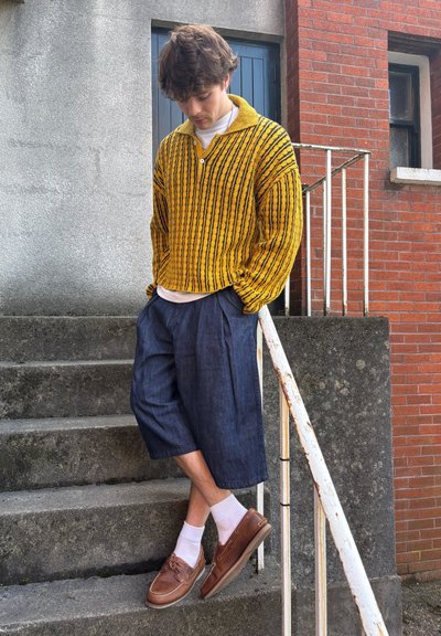 Suéter de punto a rayas amarillas y negras, pantalones cortos de mezclilla oscuros y holgados, zapatos de barco marrones y calcetines blancos, de pie en unos escalones exteriores.