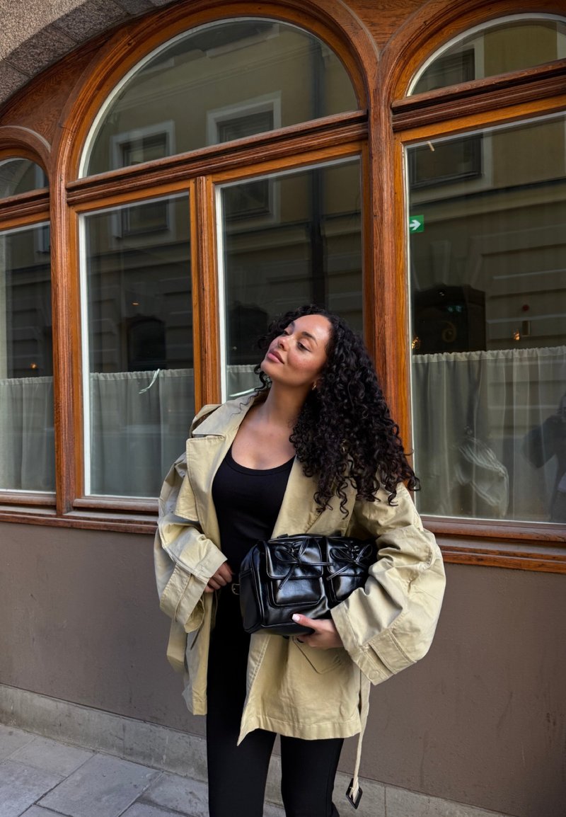 Femme aux cheveux bouclés portant une veste beige oversize et une tenue noire, tenant un sac à main en cuir noir devant des fenêtres en bois arquées.