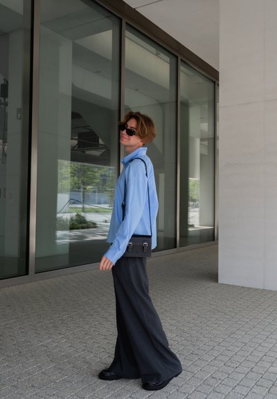Camisa de manga larga de color azul claro, pantalones anchos negros y un pequeño bolso cruzado negro con herrajes plateados. Texturas suaves, entorno urbano moderno.