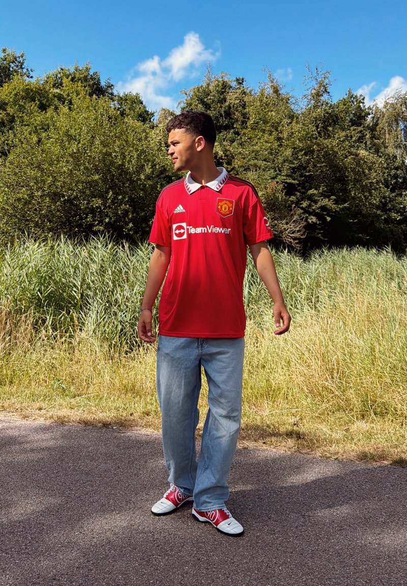Jeune homme en maillot de football rouge de Manchester United, jean bleu clair et baskets rouges, debout sur un chemin pavé près d'herbes hautes et d'arbres.