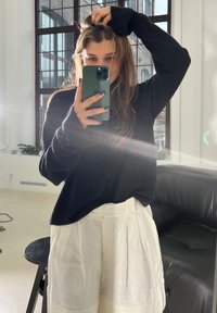 Maglione nero a maglia con maniche lunghe, abbinato a pantaloni corti bianchi con texture e pieghe. Lo sfondo include un divano in pelle nera e finestre.