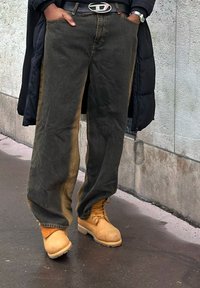 Črne široke kavbojke z teksturirano površino, nošene s svetlo rjavimi delovnimi škornji z vezalkami in grobim podplatom; pas z zaponko iz kovine.