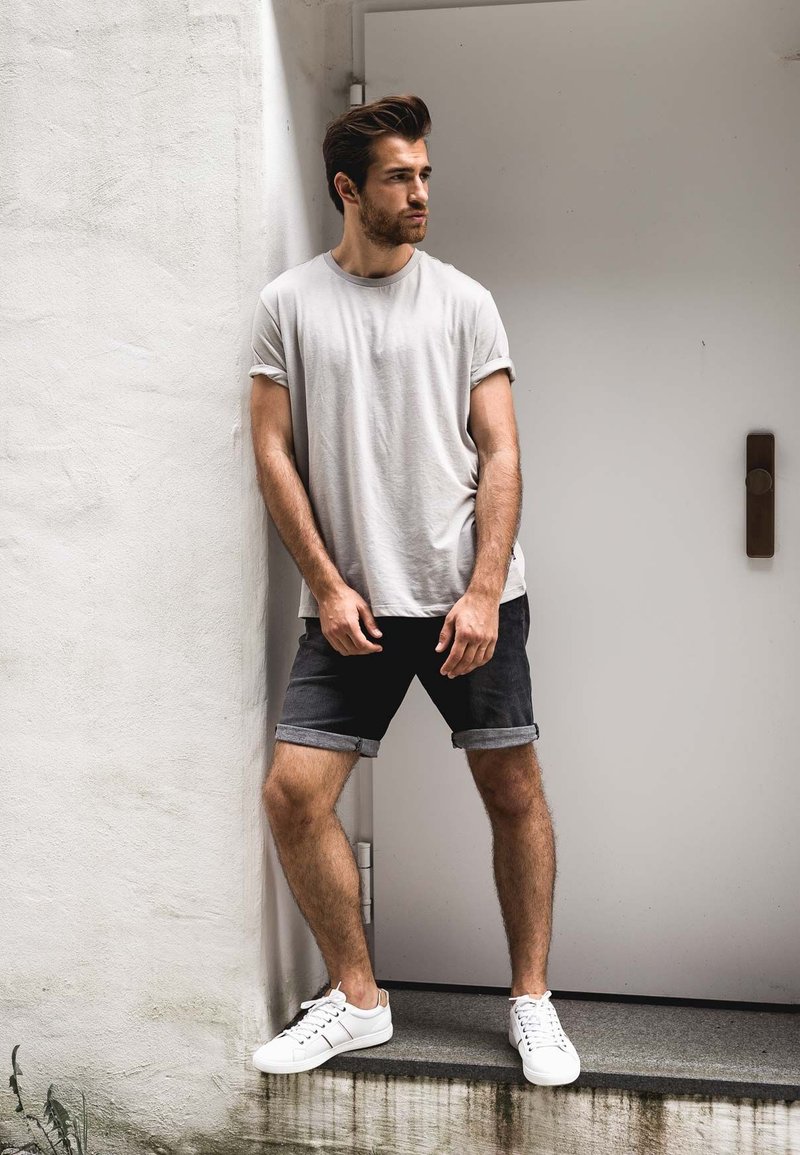 Mannen lichtgrijze crew-neck t-shirt, donkerdenimshorts met omslagen en witte sneakers. Staande pose tegen een eenvoudige witte muur en deur.