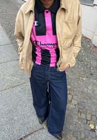 Beige jas van imitatieleren over een zwart-roze gestreept poloshirt met logo. Donkerblauwe jeans met wijde pijpen, bruine schoenen, op een geplaveide ondergrond.