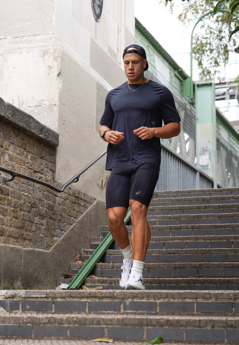 Uomo vestito con abiti sportivi neri che corre giù per le scale in pietra all'aperto vicino a un muro di mattoni e a una ringhiera metallica.