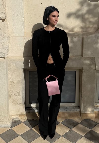 Mujer con cabello corto y oscuro, vestida con un top negro con cremallera y pantalones, sosteniendo un pequeño bolso rosa, de pie frente a una pared de piedra beige.