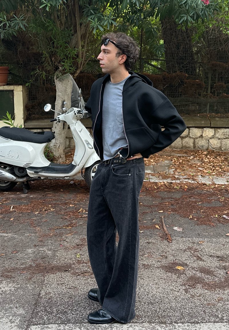Schwarze Zip-Up-Hoodie über einem hellgrauen T-Shirt, kombiniert mit lockeren schwarzen Jeans und schwarzen Schuhen. Ein weißer Roller ist teilweise im Hintergrund sichtbar.