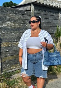 White pinstriped button-up shirt worn over a white crop top, teamed with distressed blue denim shorts and a blue tote bag featuring silver lettering.
