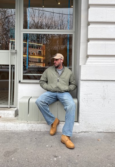 Hombre con gorra beige, chaqueta verde, vaqueros azules y botas marrones sentado en un radiador exterior junto a la ventana de una cafetería, escuchando con auriculares.