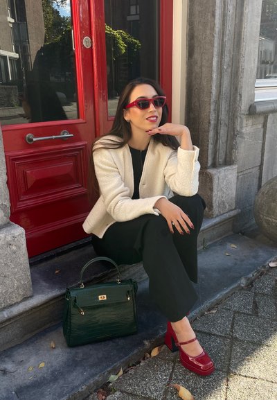 Chaqueta blanca texturizada, top negro y pantalones negros. Bolso de mano con estampado de cocodrilo verde, zapatos de plataforma rojos y gafas de sol rojas. Fondo de puerta roja.