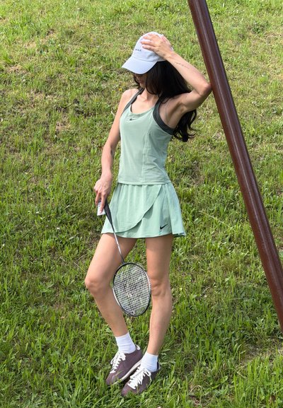 Mujer con vestido deportivo verde claro y gorra blanca que sostiene una raqueta de bádminton, de pie en un campo de hierba cerca de un poste de metal marrón.