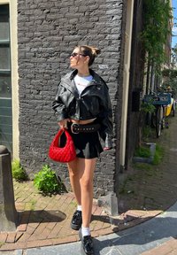 Cazaco de couro preto cropped sobre uma camisa branca, saia mini preta e sapatos de plataforma. Carrega uma bolsa tecida vermelha, em pé sobre um caminho de tijolos.