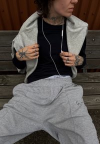 Šedé tepláky s elastickým pasem, spárované s černým tričkem s dlouhým rukávem a světle šedou zipovou bundou. Přítomné sluchátka.