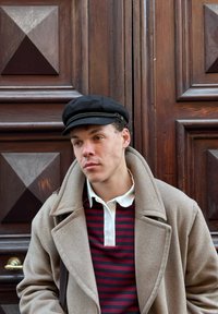 Jeune homme portant une casquette noire, un manteau beige et un polo rayé rouge et noir, appuyé contre une porte en bois foncé avec des panneaux géométriques.