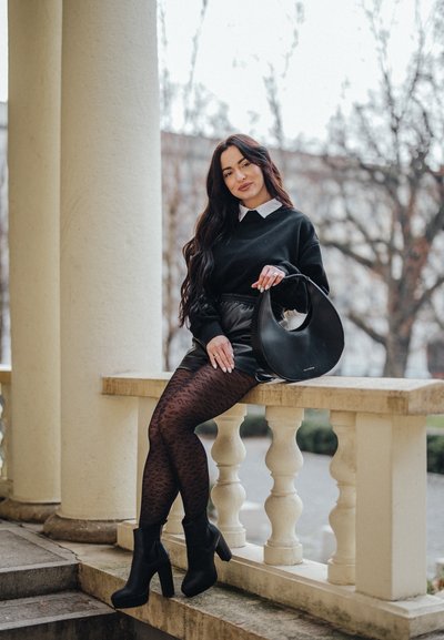 Bolso hobo negro, shorts de cuero negro, botines negros puntiagudos y medias oscuras con estampado. Suéter blanco con cuello. Escenario exterior con pilares.