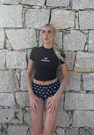 Top corto negro de manga corta con logotipo de texto rosa, combinado con pantalones cortos de bikini negros con lunares. La persona está de pie frente a una pared de piedra texturizada.