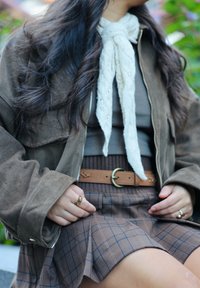 Brun ruskindsjakke med sølvlynlås, beige strikket tørklæde, brun ribbet top, brun ternet nederdel og et tanfarvet bælte med rund spænde.