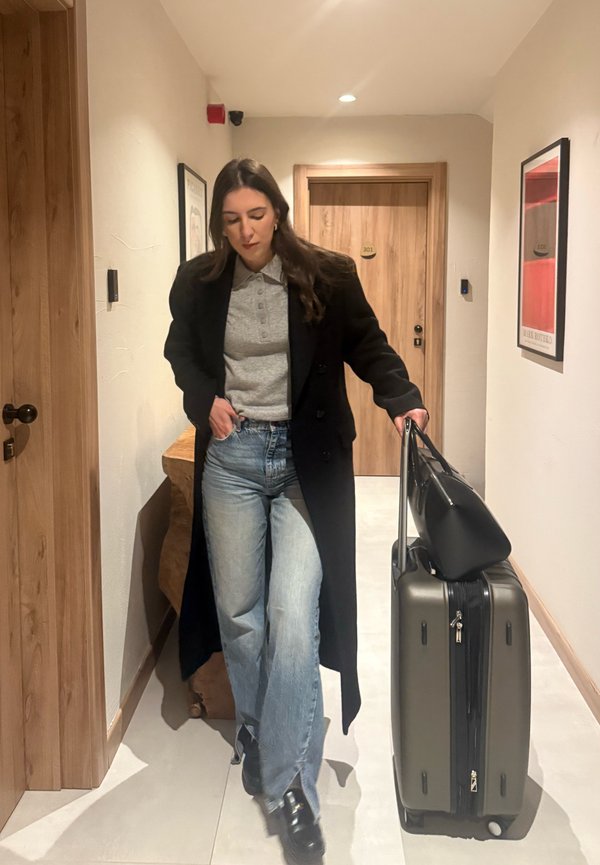 Žena v černém kabátu a džínách táhne kufr s kabelkou nahoře, kráčející chodbou hotelu s dřevěnými dveřmi a rámovaným uměním.