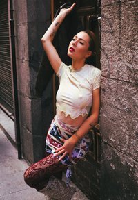 Crop top en tissu léger avec un motif abstrait, associé à une mini-jupe colorée à étages et des collants résille, sur fond de mur texturé.