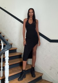 Abito nero a collarino con spacco alto alla coscia, design aderente, abbinato a stivali alla caviglia neri. Scala di legno e pareti chiare sullo sfondo.