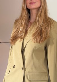 Blazer vert olive clair avec un design structuré, devant croisé à deux rangées de boutons et deux poches avant. Tissu lisse avec des détails de texture subtils.