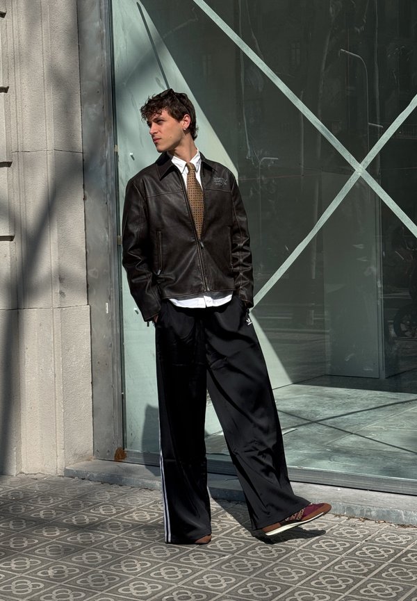 Schwarze Lederjacke mit Reißverschluss, weisses Hemd, braune gemusterte Krawatte, lockere schwarze Hose und braune Sandalen mit Riemen. Glas-Hintergrund.