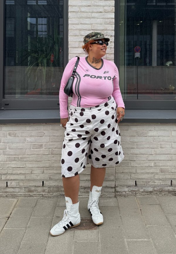 Frau trägt ein rosa Langarmshirt, weiße Shorts mit Punkten, weiße High-Top-Sneaker, Sonnenbrille und eine Camouflage-Mütze und steht an einer Ziegelwand.