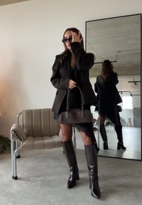 Femme en robe en dentelle noire et blazer, portant des bottes montant jusqu'aux genoux et un sac à main, ajustant ses lunettes de soleil, debout près d'une chaise et d'un grand miroir.