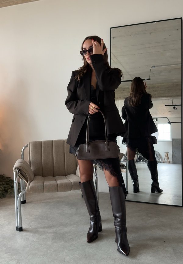 Femeie în rochie neagră din dantelă și sacou, cu cizme până la genunchi și geantă, ajustându-și ochelarii de soare, stând lângă un scaun și o oglindă mare.
