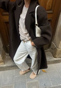 Hnědý vlněný kabát přes světle šedé tričko, bílé kalhoty a béžové huňaté pantofle; nese malou bílou tašku se zlatým popruhem.