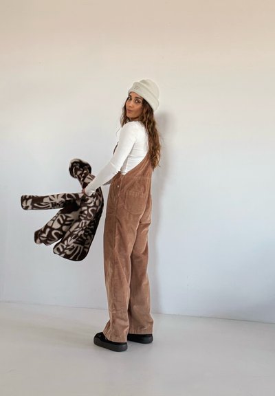 Peto de pana marrón con un corte holgado, combinado con una camiseta blanca de manga larga y un gorro beige claro. Sosteniendo un forro polar oscuro con estampado.