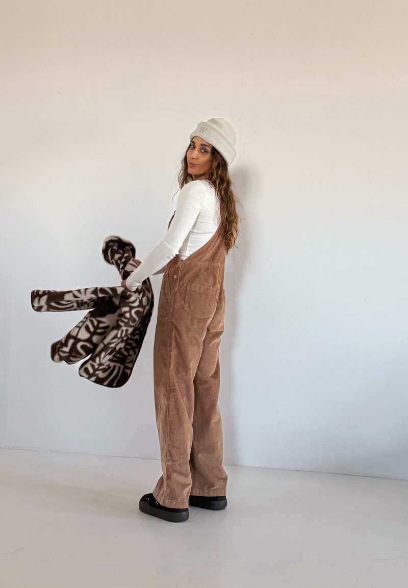 Une salopette en velours côtelé marron à coupe ample, associée à une chemise blanche à manches longues et un bonnet beige clair. Tenant une polaire foncée à motifs.