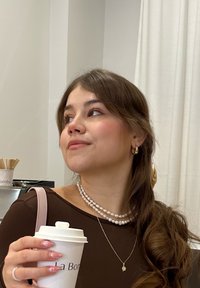 Donna che tiene in mano un bicchiere di caffè bianco con coperchio, indossando una maglietta a maniche lunghe marrone, collane di perle sovrapposte e piccoli orecchini a cerchio in oro.