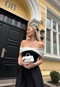 Svart kjole med hvit avskåret topp, med en sløyfe detalj. Hvit veske med håndtak. Bakgrunnen viser en svart dør og en gul vegg.