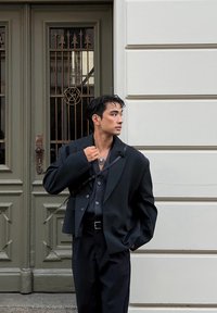 Μαύρο oversized σακάκι πάνω από ένα σκούρο πουκάμισο με κουμπιά και παντελόνι, με ασημένιο κρεμαστό κόσμημα και υφή στα μαλλιά του.