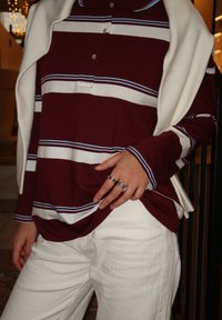 Polo a maniche lunghe a righe bordeaux e bianche con colletto, sovrapposto a un maglione color crema drappeggiato sulle spalle, abbinato a pantaloni bianchi con texture.