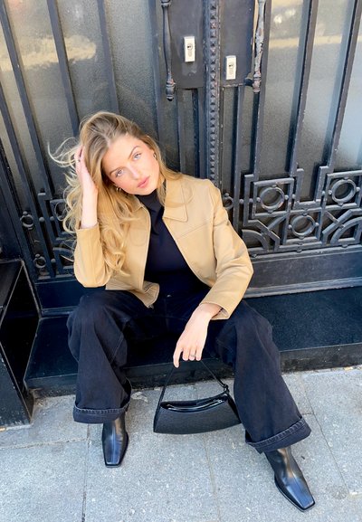 Chaqueta de cuero marrón, suéter negro de cuello alto, jeans negros de pierna ancha, botines negros de punta cuadrada y un pequeño bolso negro. El fondo presenta una puerta oscura.