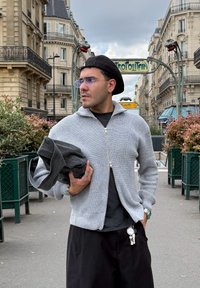 Svetlo šedý pletený kardigán na zips s rebrovanou textúrou, nosený cez čiernu košeľu. Čierny baret a tmavé nohavice dopĺňajú outfit. Drží sivú tkaninu.