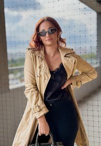 Beige Trenchcoat mit großem Kragen, schwarzes Spitzenoberteil darunter und schwarzer Rock. Die Accessoires umfassen schwarze Sonnenbrillen und eine gemusterte Handtasche.