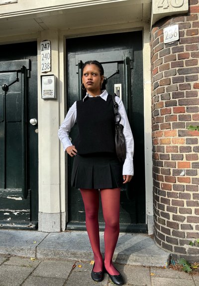 Chaleco de punto negro sobre una camiseta blanca, falda negra plisada y medias burdeos. Zapatos planos negros con detalle de lazo. De pie frente a una pared de ladrillos.