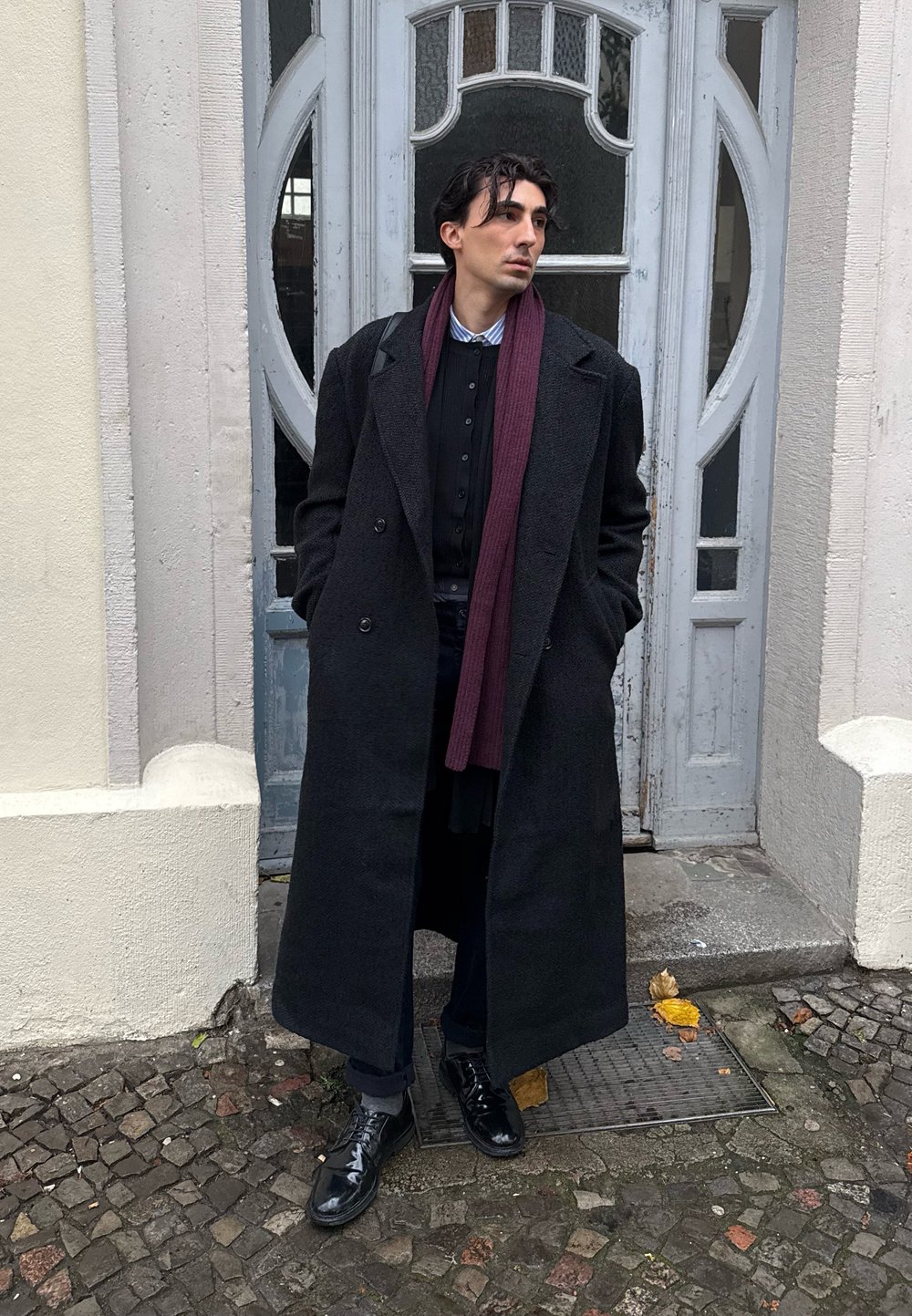 Abrigo largo negro sobre una camisa oscura, con una bufanda burdeos. Zapatos negros brillantes y pantalones oscuros sobre un fondo de adoquines y puertas grises.