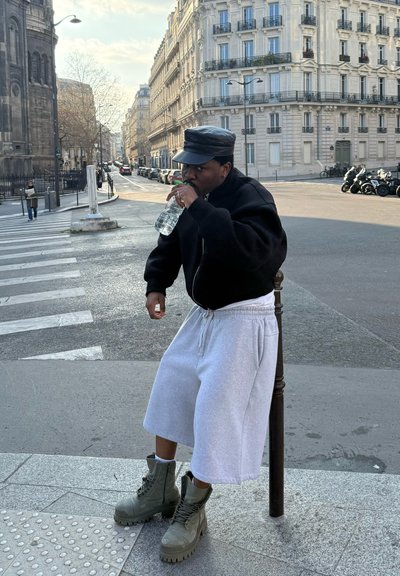 Persona con una chaqueta bomber negra, pantalones cortos deportivos grises y botas de combate verdes, sosteniendo una botella de agua en una calle de la ciudad.