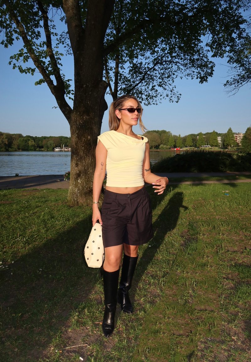 Vrouw die een lichtgele top, zwarte korte broek en zwarte kniehoge laarzen draagt, wandelt op gras nabij een boom bij een meer op een zonnige dag.