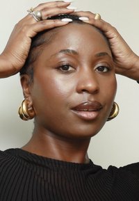Mujer con piel suave y maquillaje natural sostiene su cabello trenzado, lleva aretes de aro dorados, anillos y una camiseta negra de canalé.